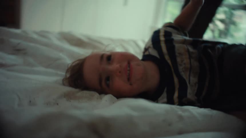 A father playfully tickles his smiling, dirt-covered son as they roughhouse together on a bed in slow motion
