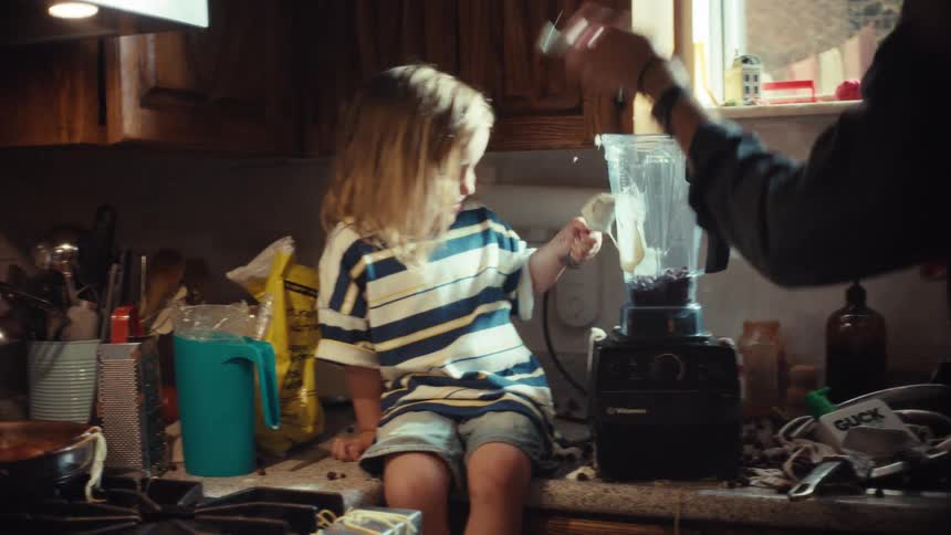 A smiling little boy sits on a messy kitchen counter helping his father add ingredients to a blender for a smoothie