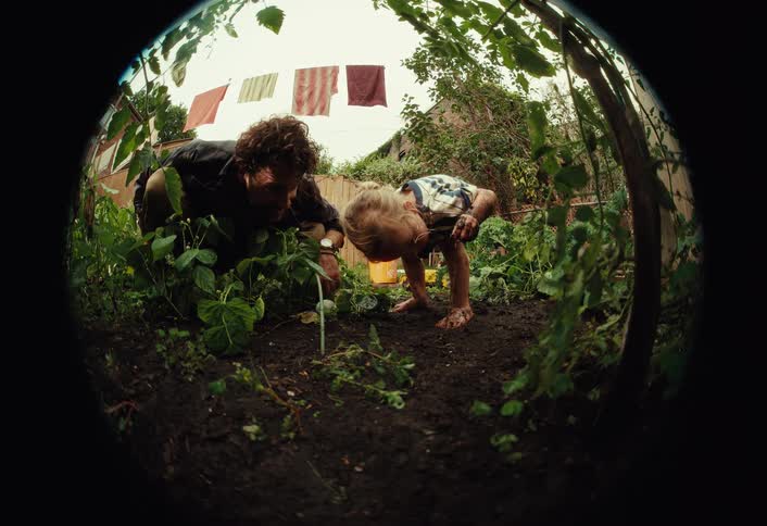 Fisheye view of a father teaching his barefoot toddler son how to garden in their muddy backyard