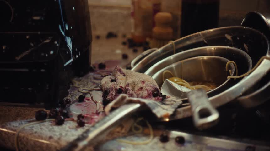 A close-up shot of a dirty and chaotic kitchen sink area, piled high with unwashed dishes and spilled food like blueberries and spaghetti