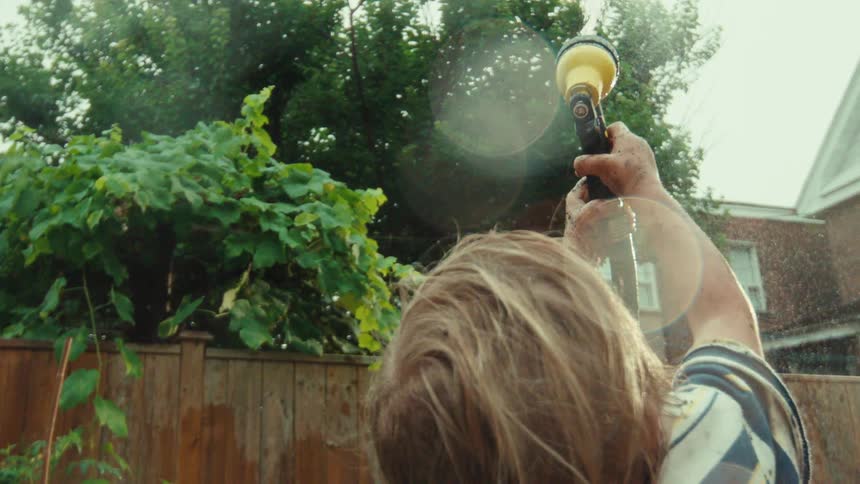 A young child with dirt-covered hands sprays a garden hose into the air on a sunny day in slow motion