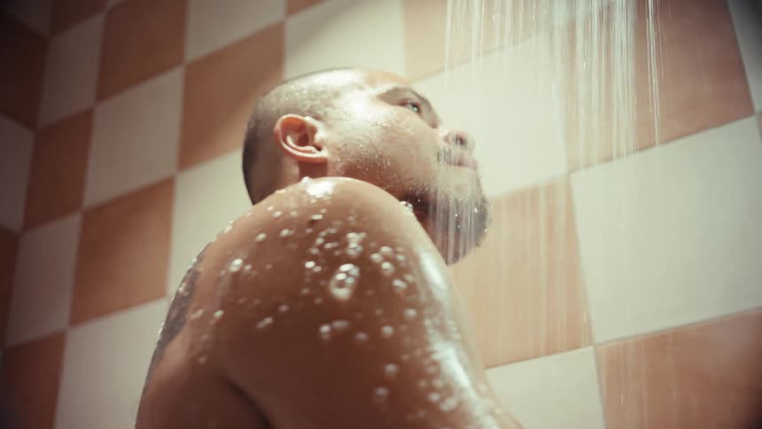 Low-angle view of a man with a tattoo lathering his shoulder with a bar of soap in a warm, tiled shower