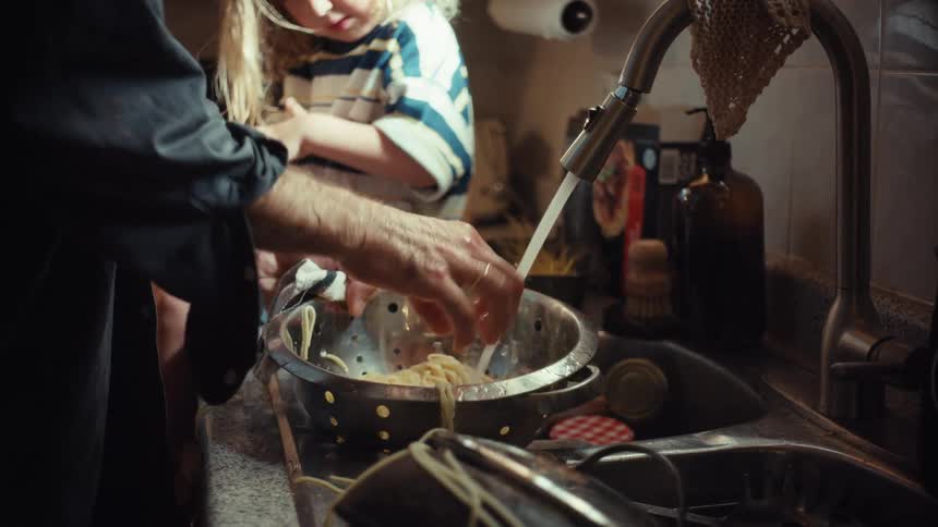 A father rinses spaghetti in a kitchen sink as his young child sits on the counter and watches