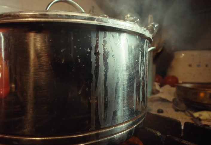 A messy stainless steel pot boils over on a gas stove, spilling liquid down the side as steam escapes from the lid