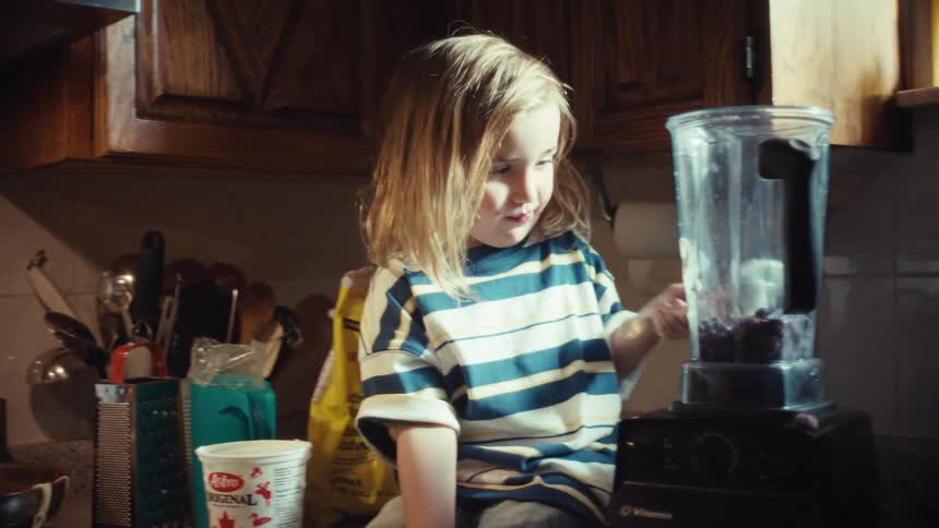 A happy child sits on the kitchen counter and helps a parent make a blueberry smoothie, adding yogurt with a spoon in slow motion