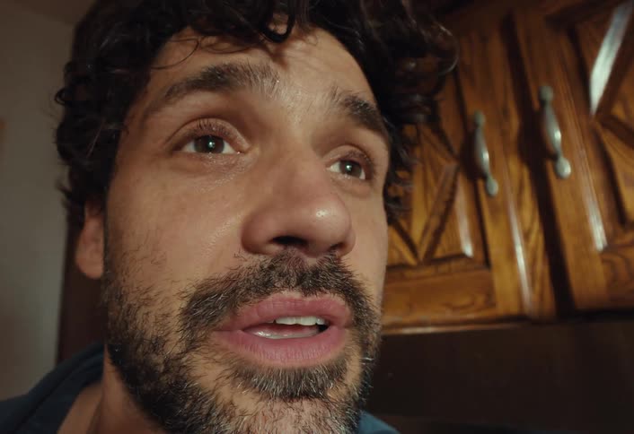 A shaky, extreme close-up shot captures a man with curly hair and a beard as he sneezes powerfully
