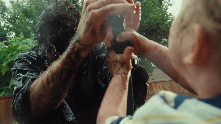A girl with muddy hands playfully sprays her father with a garden hose in the backyard in super slow motion