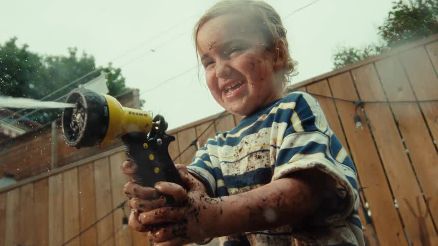 A low-angle slow-motion shot captures a gleeful toddler covered in mud spraying a garden hose in the backyard