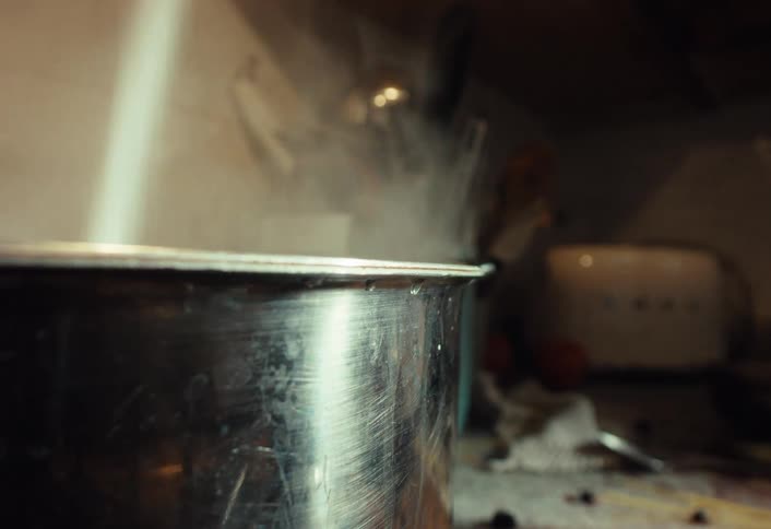 Lifting the lid from a steaming stainless steel pot on a stove and adding ingredients while cooking in a messy kitchen