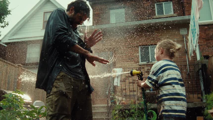 A joyful toddler boy playfully sprays his father with a garden hose in the backyard in cinematic slow motion