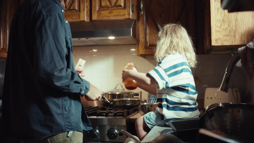 A young child sits on the kitchen counter and helps their smiling father cook dinner, pouring tomato sauce into a pan on the stove