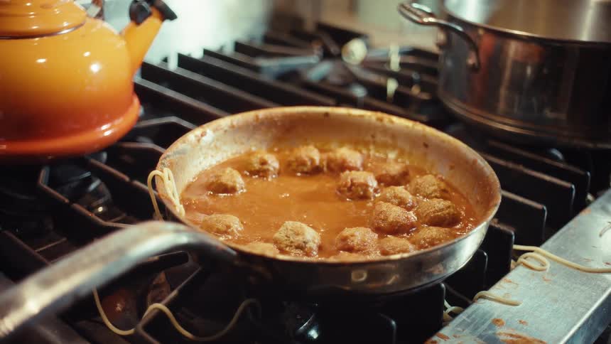 Spaghetti noodles fall from the sky into a simmering pan of meatballs and sauce on a stove