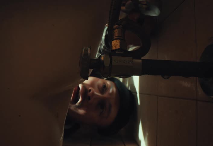 Vertical slow-motion shot of a plumber getting sprayed in the mouth by a bursting pipe while working under a toilet