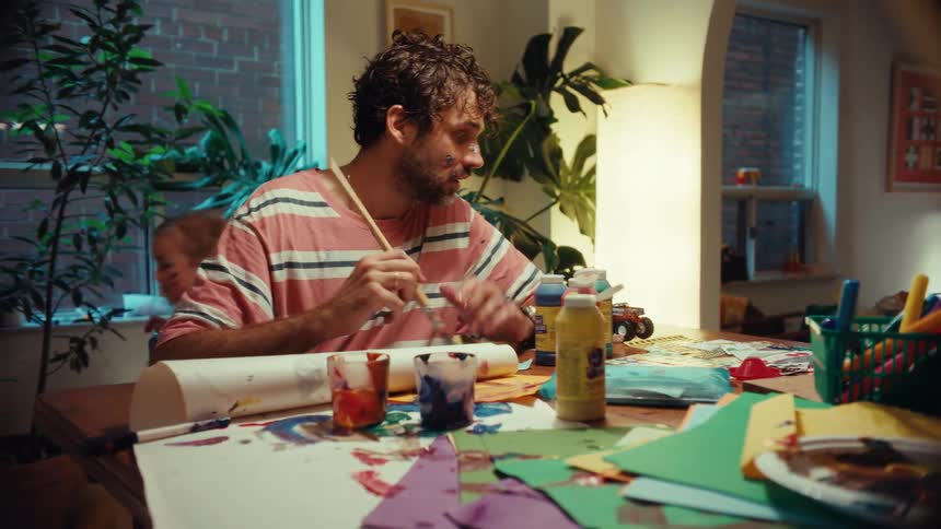 A father with paint on his face focuses on an art project at a messy table while his young child plays in the background