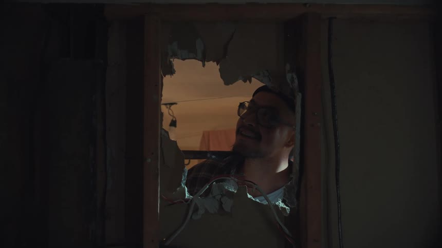 A man wearing glasses and a beanie smiles as he looks through a hole in a wall during a home renovation project
