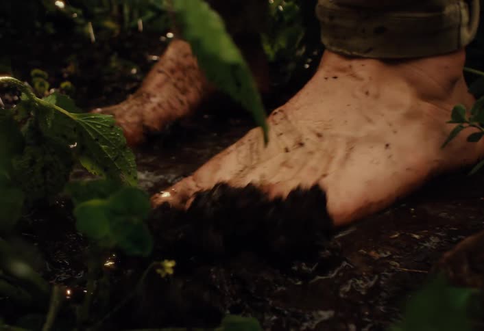 A sensory close-up shot captures bare feet squishing through wet mud as water pours down, creating a deep connection with the earth
