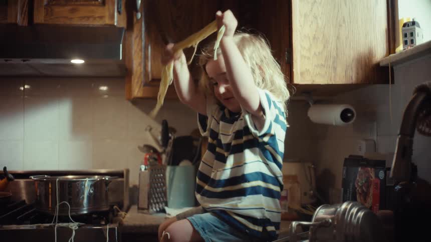A joyful little boy with long blonde hair sits on the kitchen counter, making a mess and playing with spaghetti while his father is below