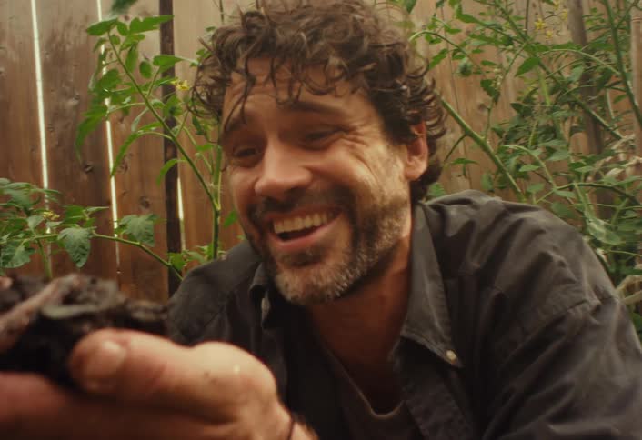 A father reacts with joyful wonder as his child places a handful of rich soil and earthworms into his open palm while gardening