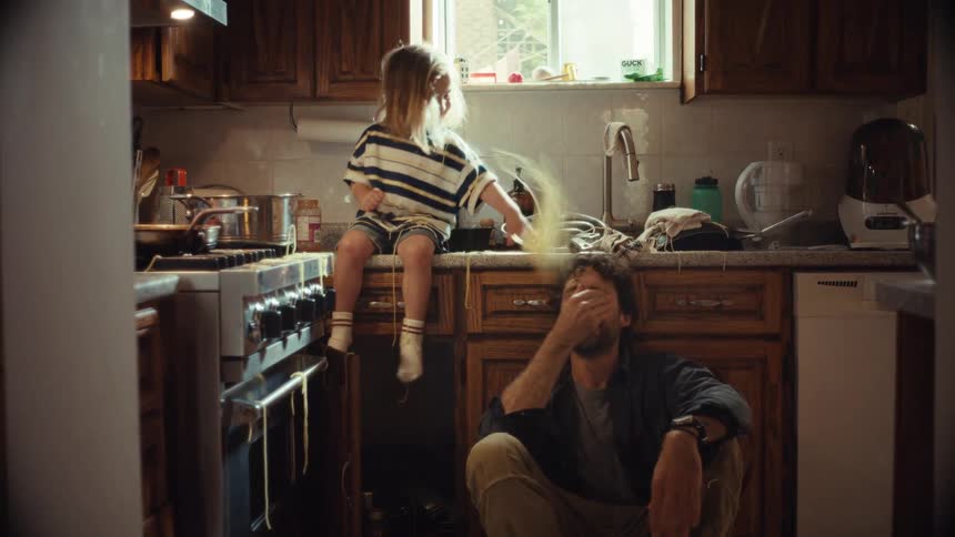 A playful father sits on the floor of a messy kitchen and makes his young child laugh by pretending to eat a handful of dry spaghetti