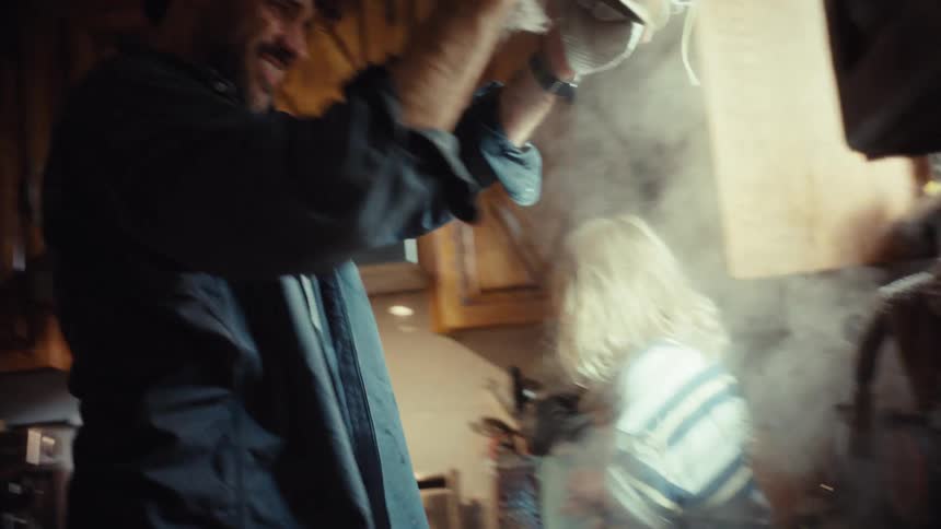 A father playfully drains hot steaming pasta into a colander as his young son watches from the kitchen counter