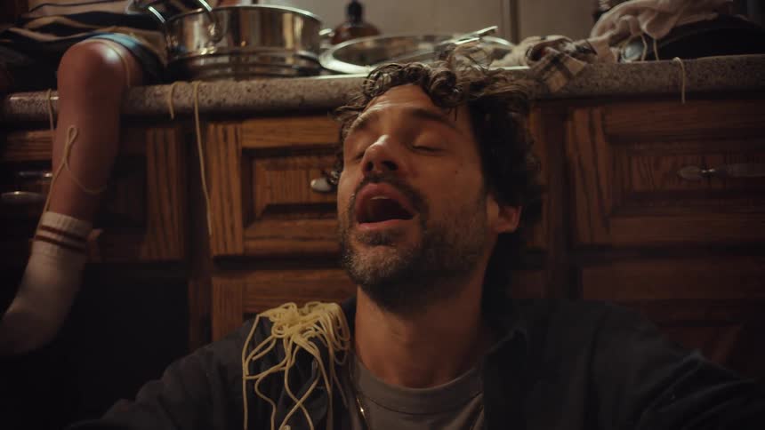 Slow motion close-up of an exhausted father having a mental breakdown as his child pours spaghetti on his head in a chaotic kitchen