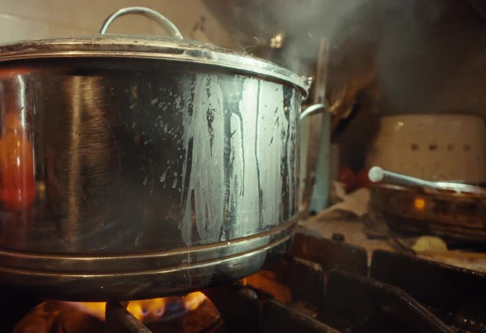 A close-up of a stainless steel pot vigorously boiling over on a gas stove as steam billows out
