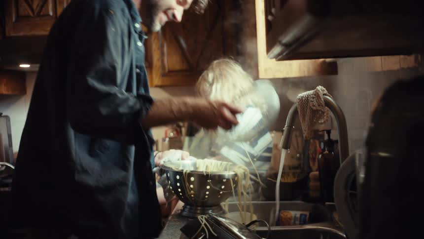 A father and his young daughter laugh together as he drains hot, steaming spaghetti in the kitchen sink
