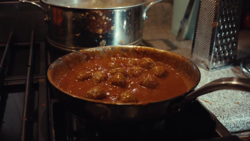 A close-up of a chef tossing meatballs in a pan of simmering tomato sauce on a gas stove