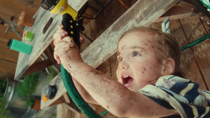 Low angle slow motion cinemagraph of a happy young girl covered in mud spraying a garden hose in the backyard