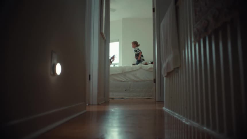A playful toddler energetically jumps on a bed with their father, viewed from a low angle down a dimly lit hallway