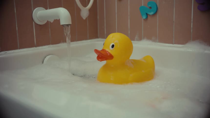 A classic yellow rubber duck floats in a soapy bubble bath while the water runs