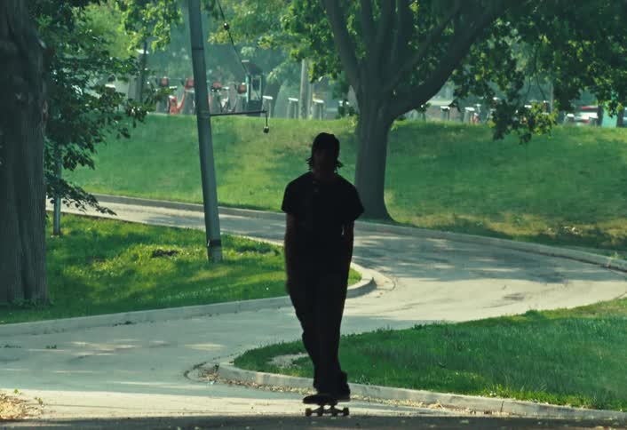 A young skateboarder cruises down a winding park path in slow motion on a sunny day