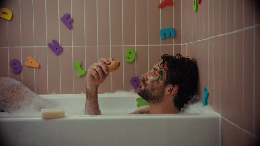 A quirky man with a painted face blissfully eats a hamburger while taking a bubble bath in a children's bathroom