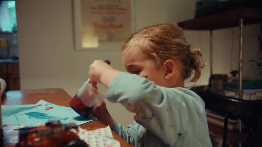 Playful toddler with paint on their face mixes colors while their happy father watches them at the kitchen table