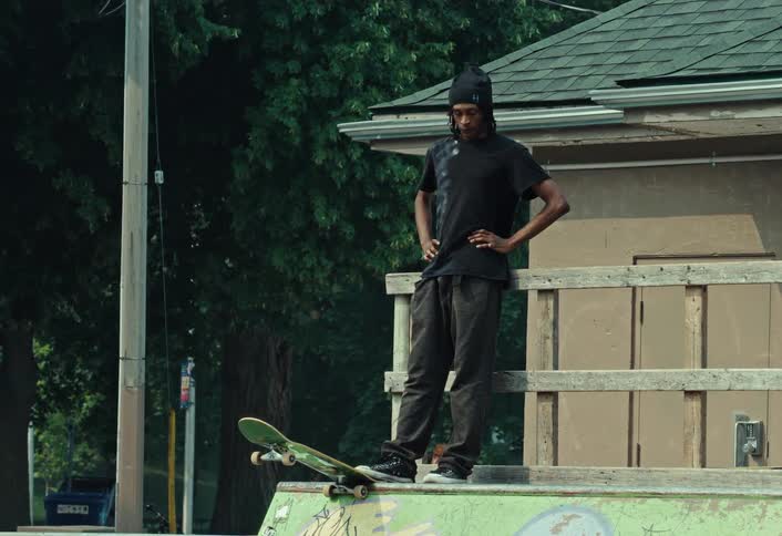 A skateboarder with dreadlocks and a beanie stands with his hands on his hips, contemplating his next move at the top of a skatepark ramp