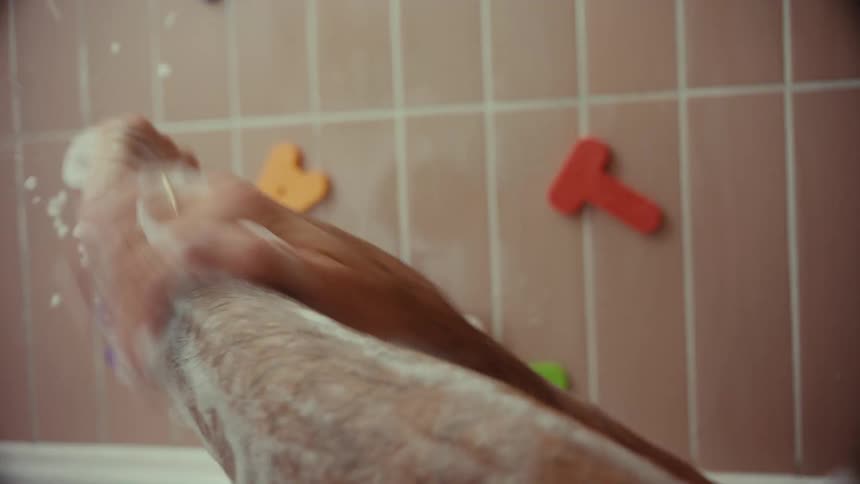 A first-person view of a man lathering his arms with soap while taking a bath in a tub with children's toys on the tiled wall