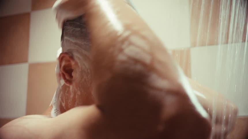 An intimate close-up shows a man with a shaved head thoroughly washing his face and scalp with soap in the shower