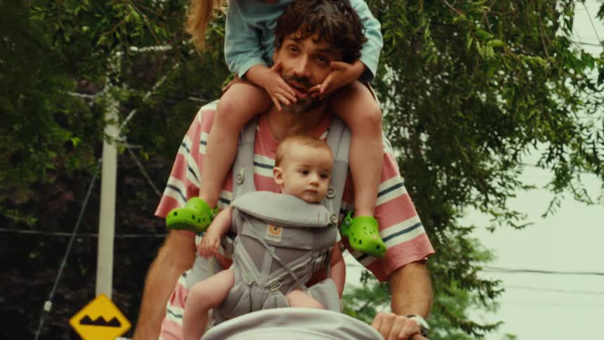 A tired father walks down a suburban street with his toddler daughter on his shoulders and his baby in a carrier while pushing a stroller