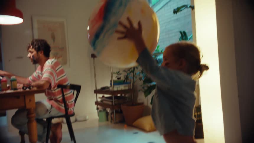 A barefoot toddler chases a large beach ball around a messy room while her father paints at the dining table
