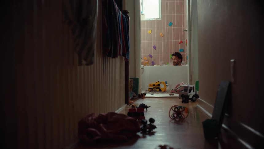 A father relaxes in a bubble bath at home, surrounded by kids' toys, before his child closes the bathroom door