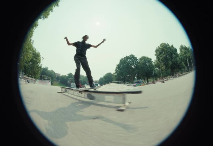 A slow-motion fisheye view captures a skateboarder as he attempts to grind a rail, ultimately losing his balance and falling at an outdoor skatepark