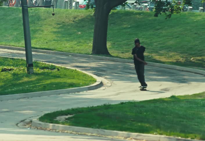 A young man skateboards down a winding paved path through a lush green park on a sunny day