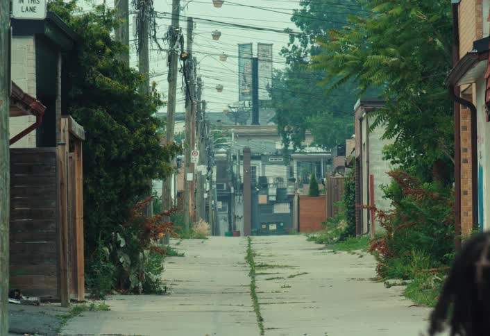 A person with dreadlocks walks away from the camera down a long, overgrown urban alleyway on a cloudy day