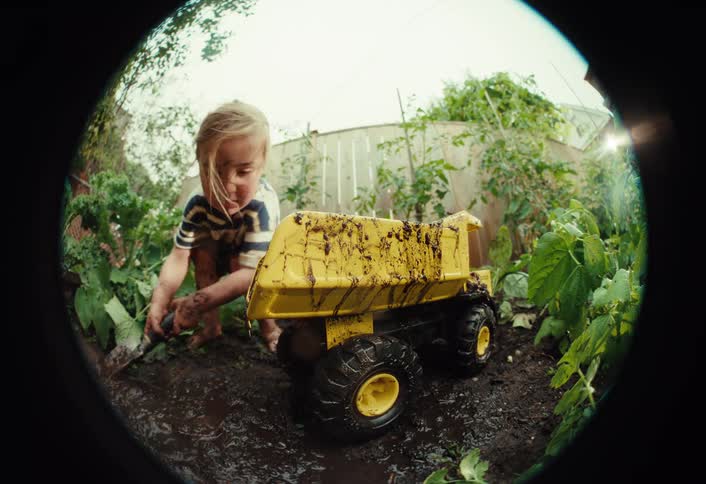 Fisheye view of a barefoot toddler getting messy while pouring mud from a bucket and playing with a toy dump truck in a backyard garden