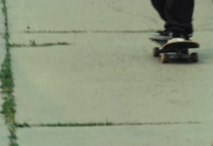 Vintage film footage shows a close-up of a person's legs and skateboard riding on a cracked concrete sidewalk