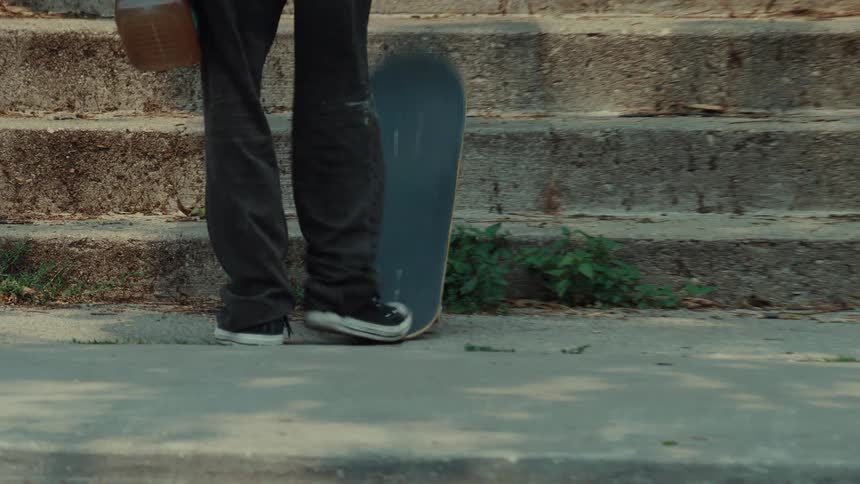 A skateboarder rides along a city sidewalk, stops, picks up their board, and walks up a flight of stairs
