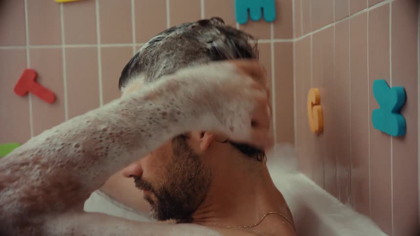 A bearded man lathers his hair with shampoo while relaxing in a bubble bath, accompanied by a yellow rubber duck