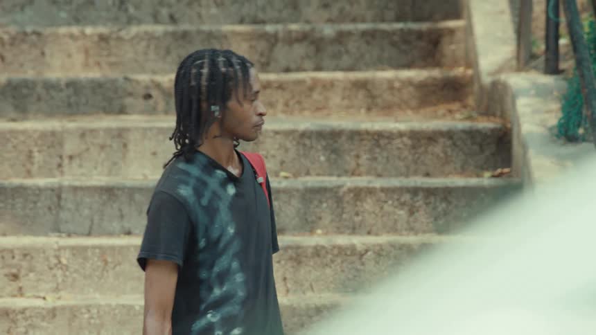 A young man with dreadlocks and a red backpack stands with his skateboard by concrete stairs before walking away
