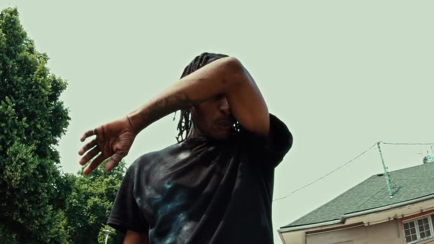 A young Black man with dreadlocks shields his eyes from the sun and wipes away sweat in slow motion on a hot summer day