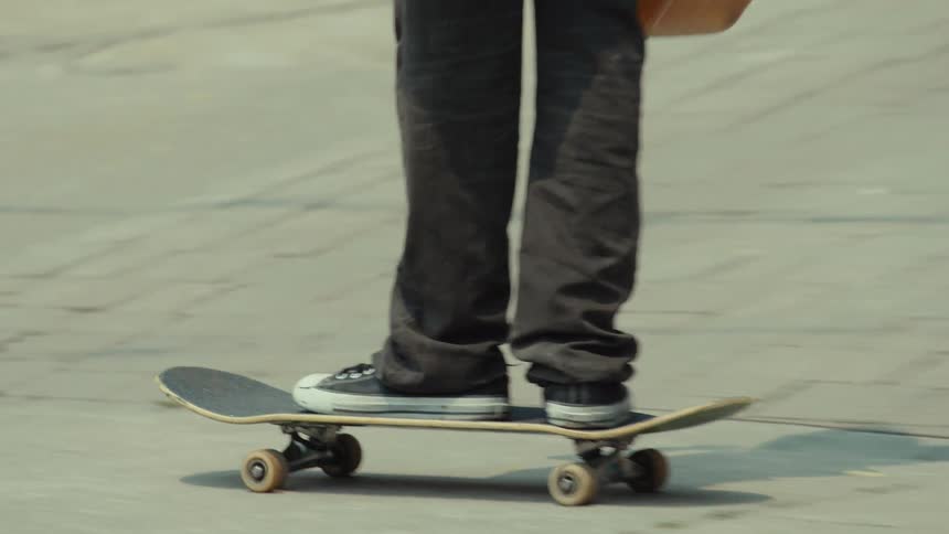 A person wearing a red backpack skateboards along a city sidewalk, passing traffic before performing a trick and walking out of frame